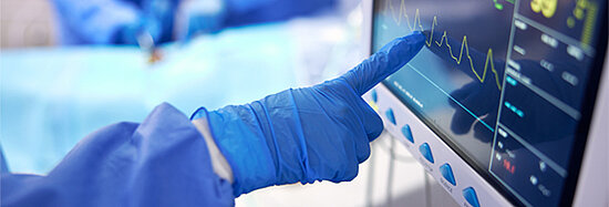 Foto, das einen behandschuhten Finger zeigt, der auf den Monitor eines EKG in einem Operationssaal drückt.