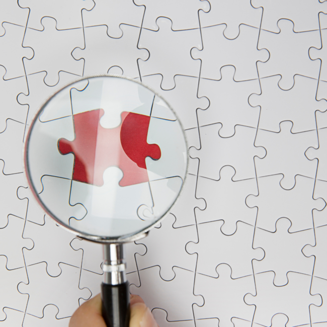ein weißes Puzzle, eine Lupe zeigt auf ein fehlendes Puzzle-Teil.