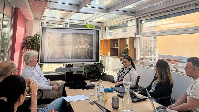 ein Foto von einem Meetingraum mit Mitarbeiter*innen der snap, auf einem großen Bildschirm sieht man noch zusätzliche Mitarbeiter*innen über Teams
