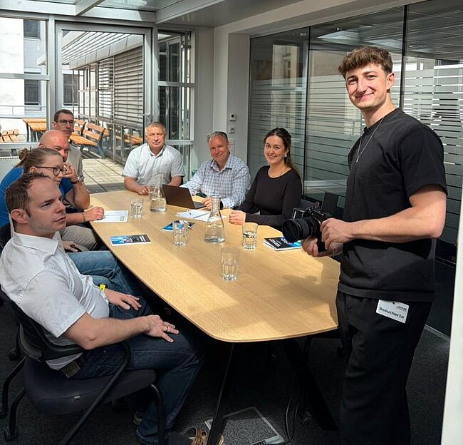 Ein Foto von unseren Mitarbeiter*innen wie sie um einen Meetingtisch sitzen, davor stehend ein Fotograf der in die Kamera lächelt