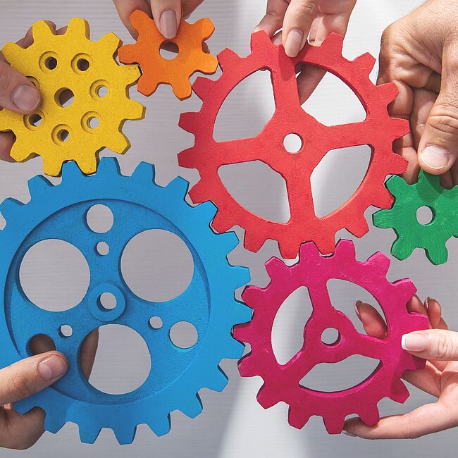 mehrere Hände die bunte Zahnräder zusammenhalten